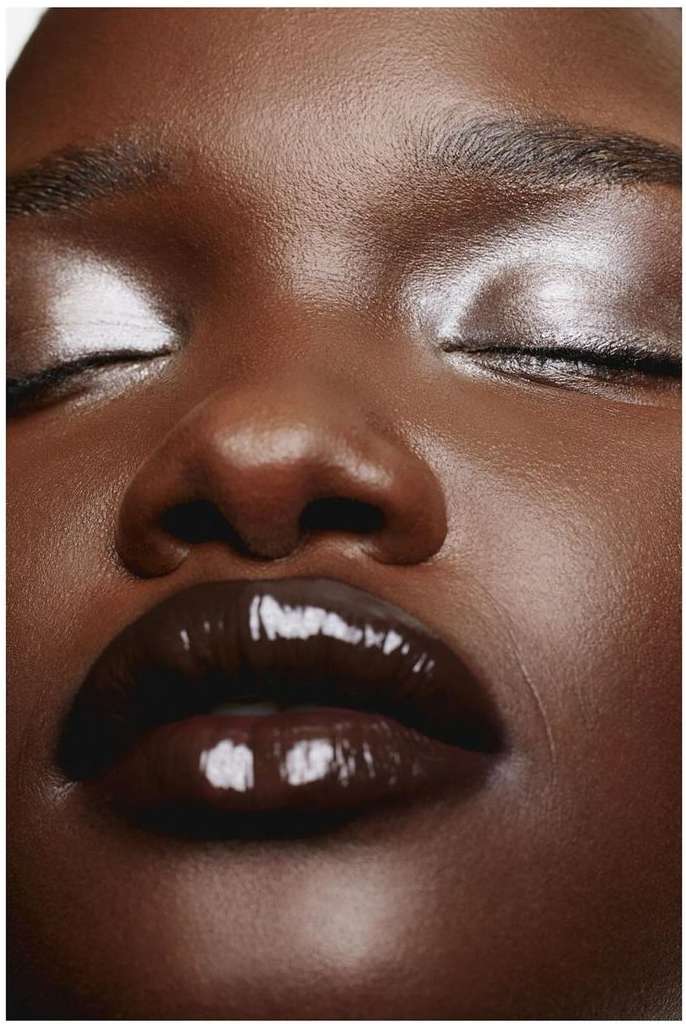 Close-up portrait of a dark-skinned woman with closed eyes, glossy dark brown lips, and shimmering silver eyeshadow. Beauty shot.