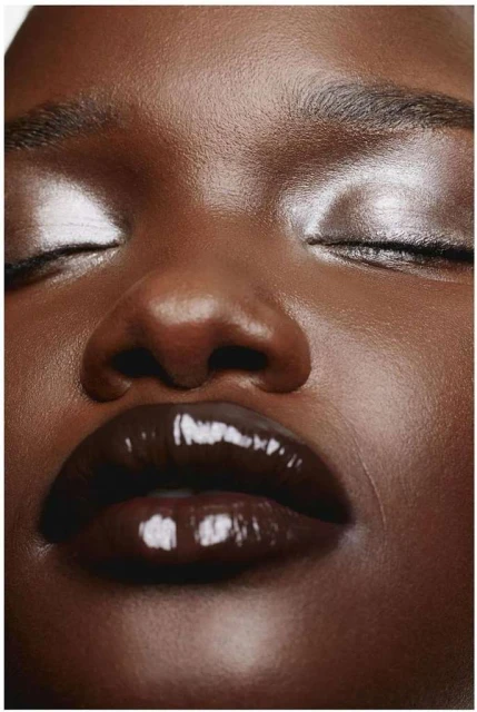 Close-up portrait of a dark-skinned woman with closed eyes, glossy dark brown lips, and shimmering silver eyeshadow. Beauty shot.