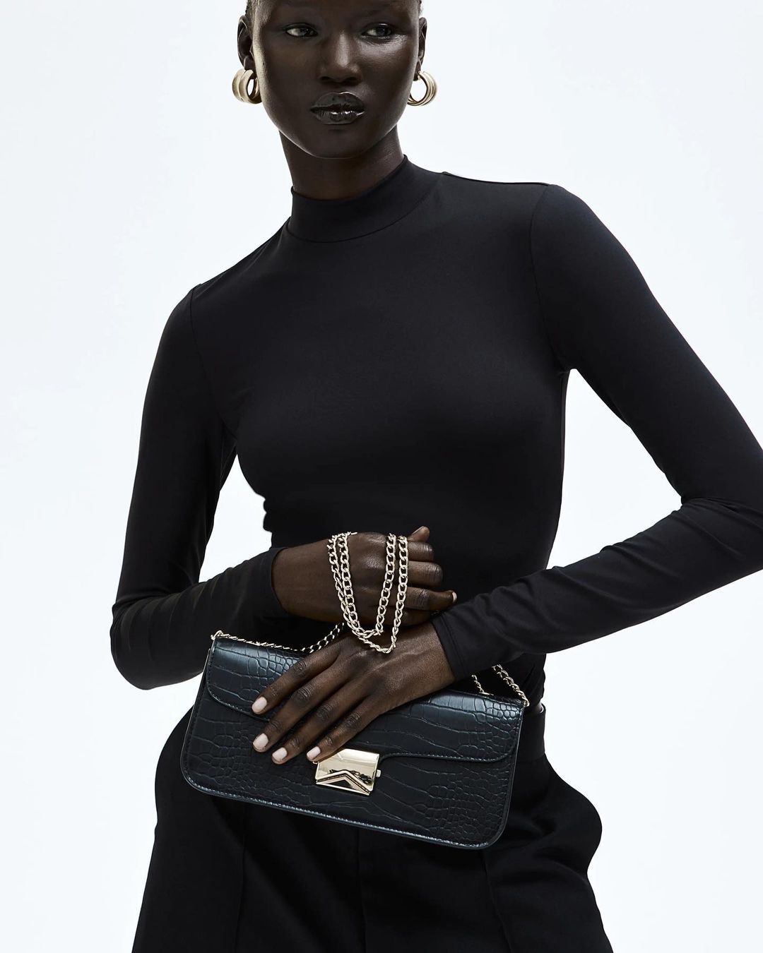 Dark-skinned woman in a black turtleneck holding a black faux crocodile purse with a gold chain and clasp, studio shot.