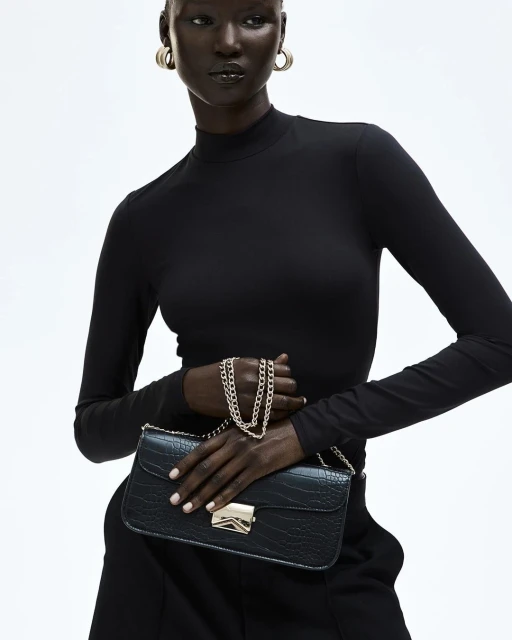 Dark-skinned woman in a black turtleneck holding a black faux crocodile purse with a gold chain and clasp, studio shot.