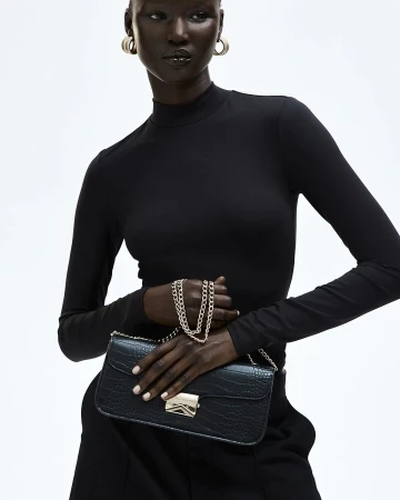 Dark-skinned woman in a black turtleneck holding a black faux crocodile purse with a gold chain and clasp, studio shot.