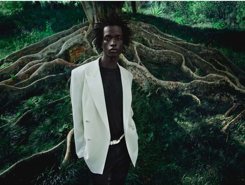 Portrait of an adult male wearing a light-colored blazer and dark pants, posing outdoors with a large tree root in the background.