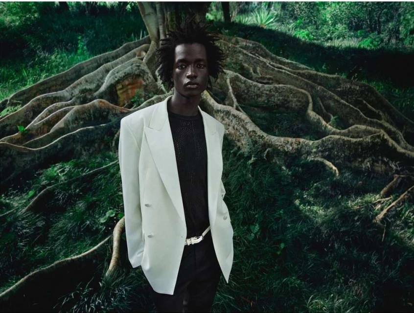 Portrait of an adult male wearing a light-colored blazer and dark pants, posing outdoors with a large tree root in the background.