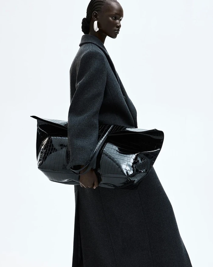 Black woman wearing a dark grey wool coat and holding a large black patent leather handbag against a white background