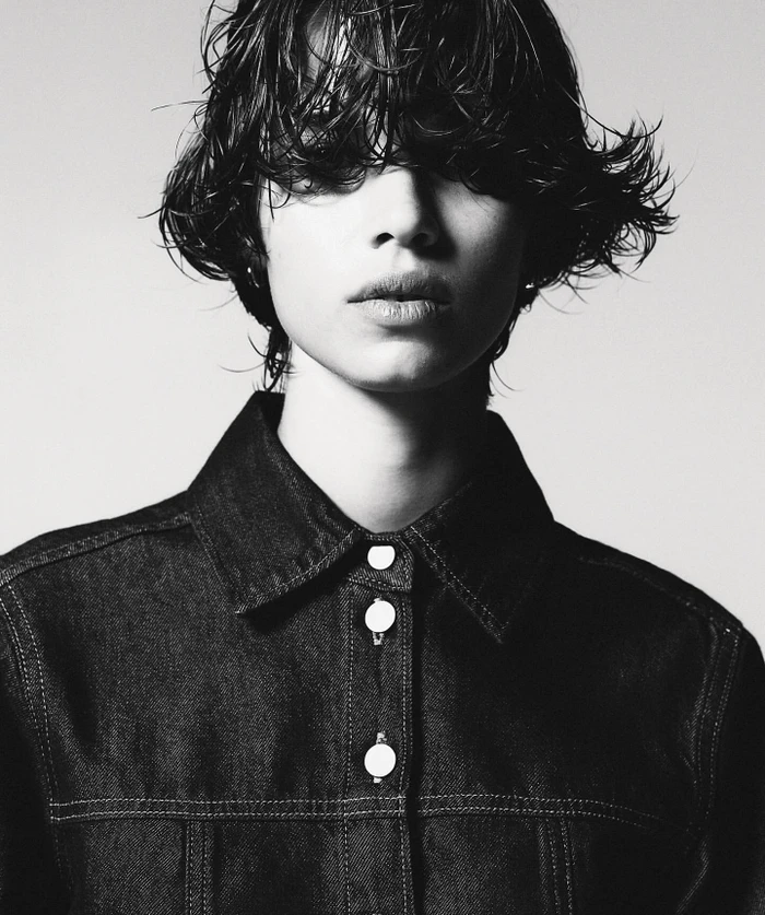 Close-up monochrome portrait of a person with dark wet hair wearing a dark denim jacket against a light background.