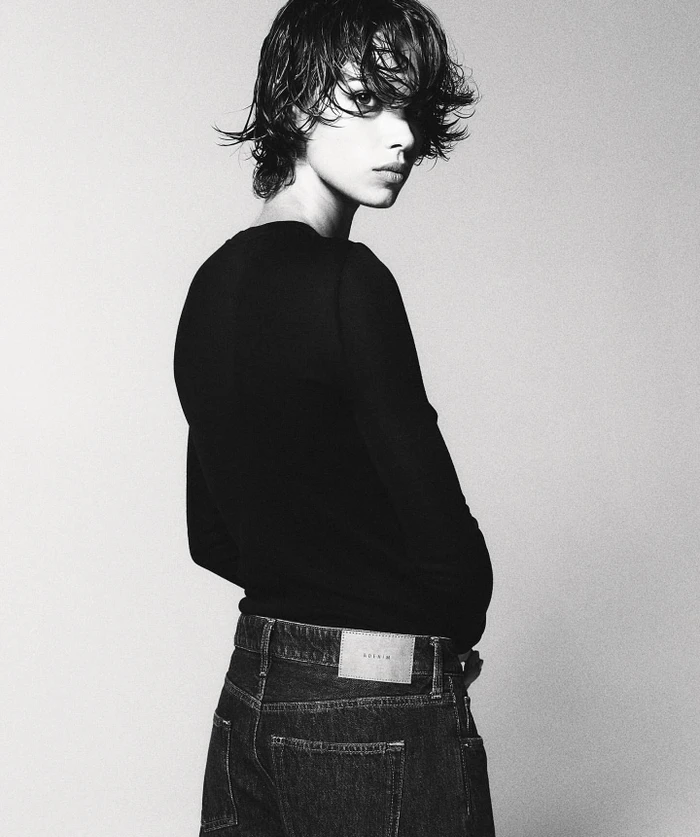 Model with wet hair wearing a black long-sleeve top and dark denim jeans against a seamless white background