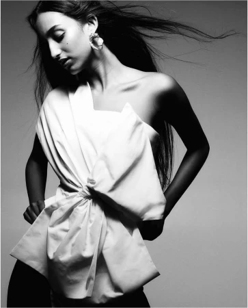 Black and white portrait of a woman with long dark hair wearing a sculptural white top and large earring against a grey backdrop.