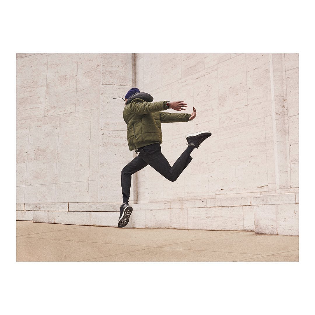 An adult male joyfully leaps against a stone wall, wearing a puffer jacket, dark pants and shoes, and a knit cap, in an outdoor setting.