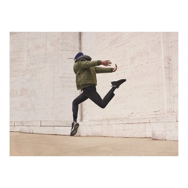 An adult male joyfully leaps against a stone wall, wearing a puffer jacket, dark pants and shoes, and a knit cap, in an outdoor setting.