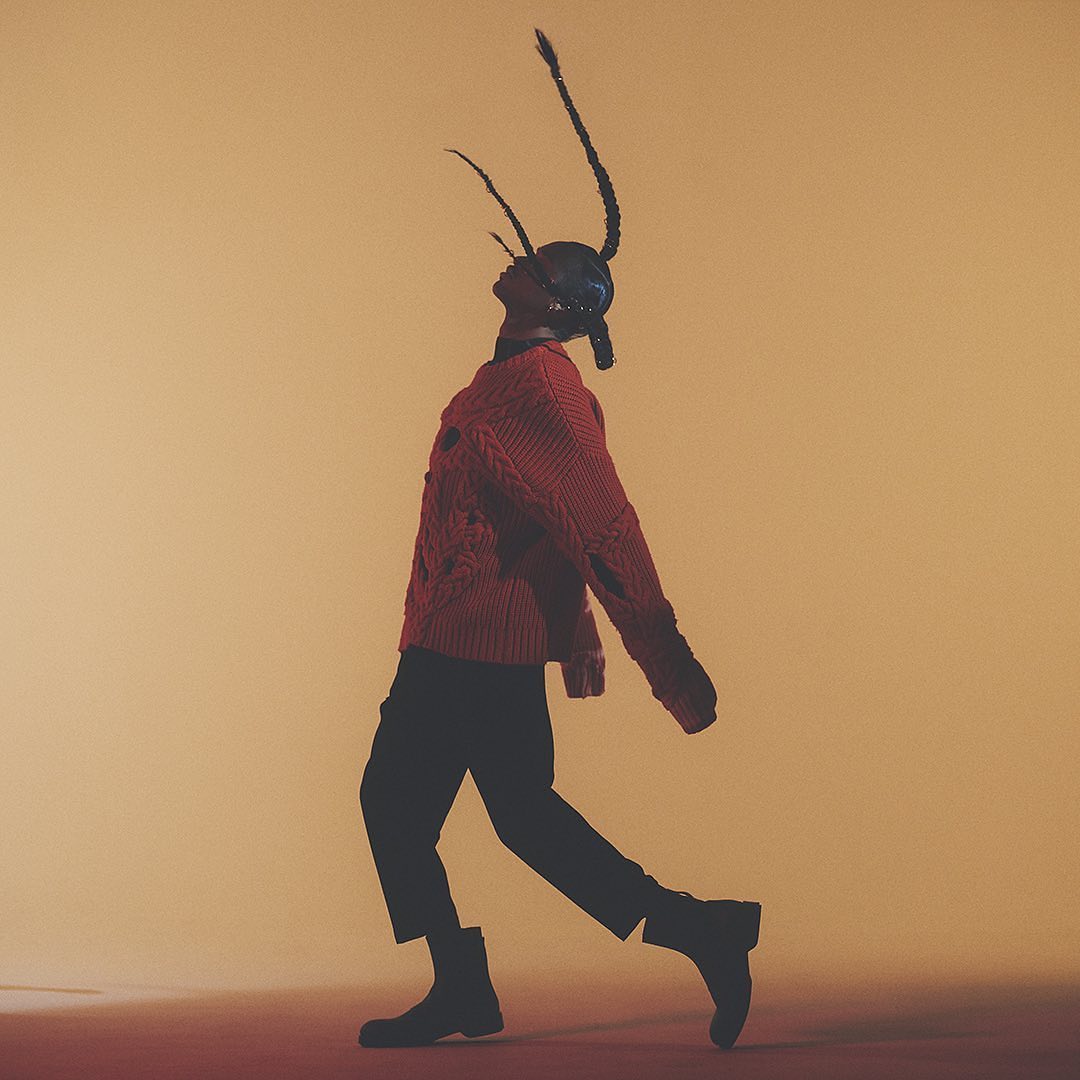 Full-length shot of a person with braided hair wearing a red sweater, black pants, and boots, walking against a gradient yellow background.