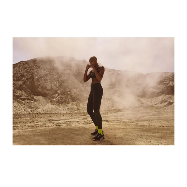 Athletic woman in black sportswear stands in boxing stance on a sandy surface with a light tan dune and hazy sky in the background.