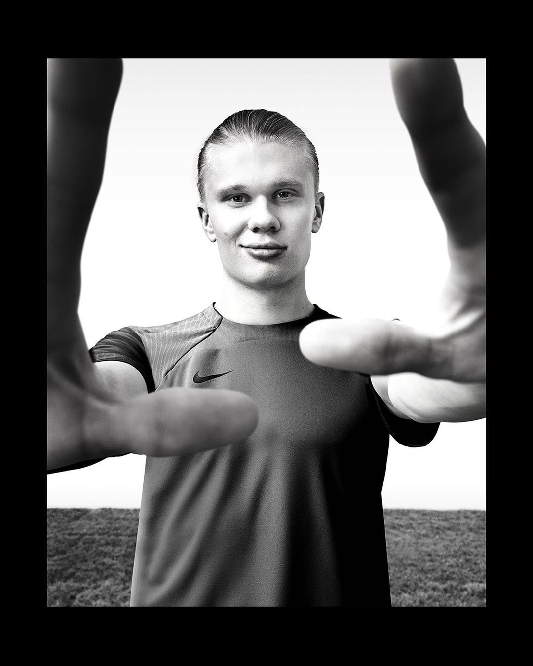 Close-up black and white portrait of a smiling athlete with slicked-back hair, framed by two hands, against a bright, blurred background.