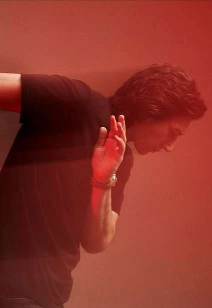 Man in black t-shirt leans near red wall with hand raised, wearing watch, captured in soft lighting, creating a simple yet striking composition.