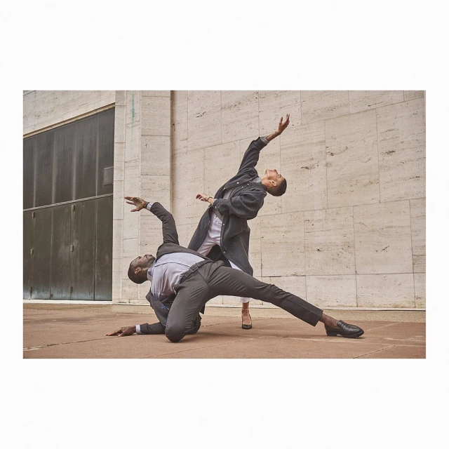 Two people in suits pose dynamically outdoors in an urban setting, one leaning back, the other balancing on one leg, against a stone wall.