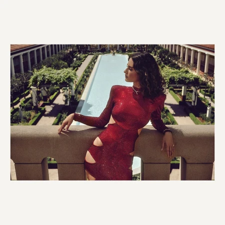 A woman in a sparkly red cut-out dress leans on a stone balcony overlooking a formal garden with a long reflecting pool.