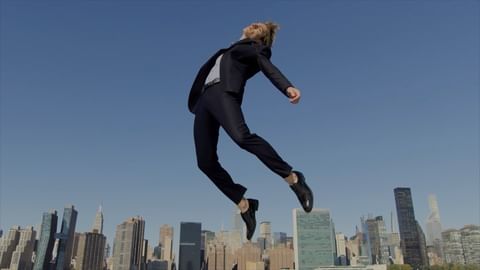 Man in a suit jumps in front of a city skyline against a clear blue sky, creating a dynamic and striking visual contrast.