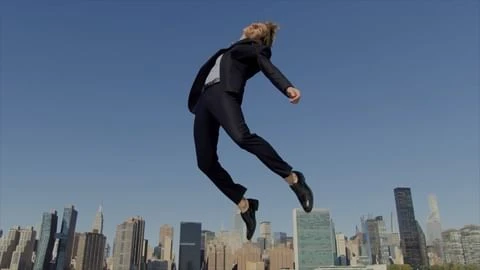 Man in a suit jumps in front of a city skyline against a clear blue sky, creating a dynamic and striking visual contrast.
