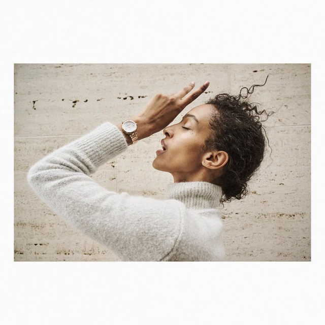 Woman in a grey sweater with a gold watch posing in front of a stone wall. The shot captures beauty and fashion with natural light.