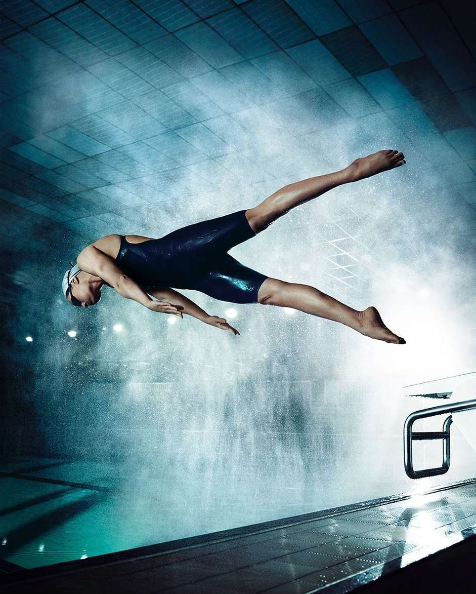 Female athlete dives into a swimming pool in a blue swimsuit. The water is clear with bright lights reflecting off the surface.