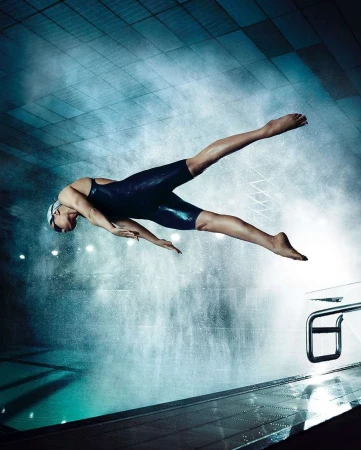Female athlete dives into a swimming pool in a blue swimsuit. The water is clear with bright lights reflecting off the surface.