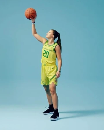 Female basketball player in action, wearing a yellow and green jersey, holding a ball against a blue background in a full length shot.