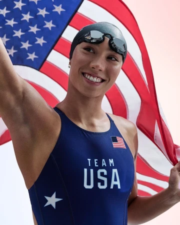 Smiling female swimmer in Team USA swimsuit and cap with goggles holding an American flag against a light-colored background.