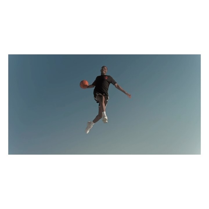 A man in athletic wear jumps with a basketball against a blue sky in an eye-level, medium shot. The image is brightly lit.