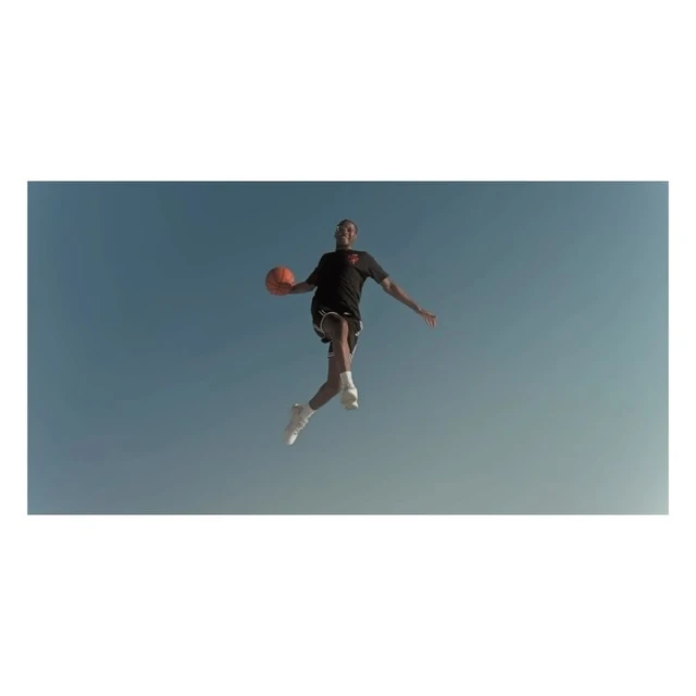 A man in athletic wear jumps with a basketball against a blue sky in an eye-level, medium shot. The image is brightly lit.
