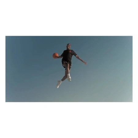 A man in athletic wear jumps with a basketball against a blue sky in an eye-level, medium shot. The image is brightly lit.