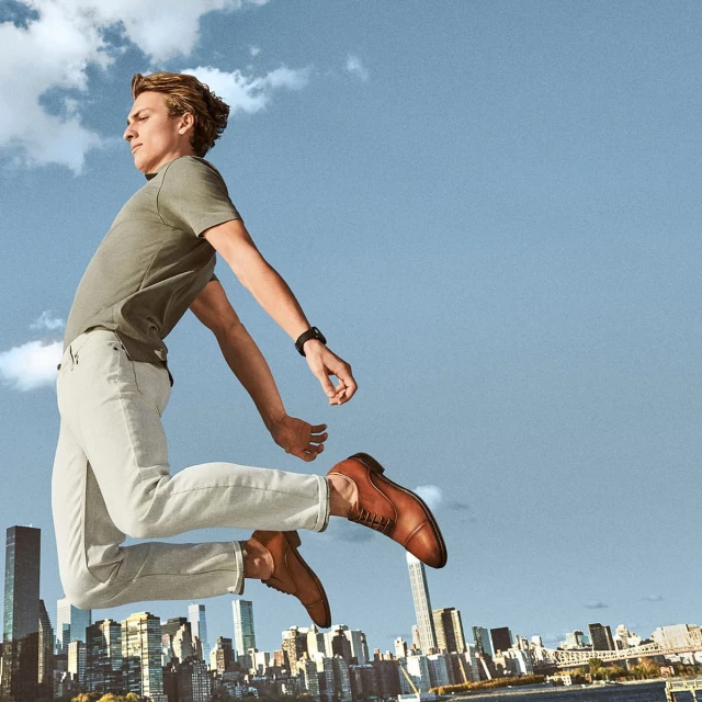 Young adult male mid-air in a leaping pose with a cityscape backdrop, wearing casual clothes, brown leather dress shoes, and a smartwatch.
