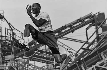 Athletic Black man leaps high outdoors in a monochromatic photo, wearing shorts and sneakers against an industrial background under daylight.