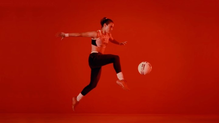 Athletic woman in sports bra and leggings practices soccer kick in an indoor studio with a plain red background and dynamic lighting.