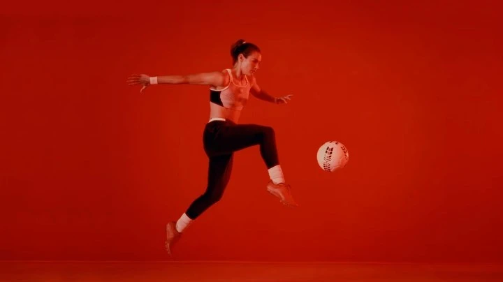 Athletic woman in sports bra and leggings practices soccer kick in an indoor studio with a plain red background and dynamic lighting.