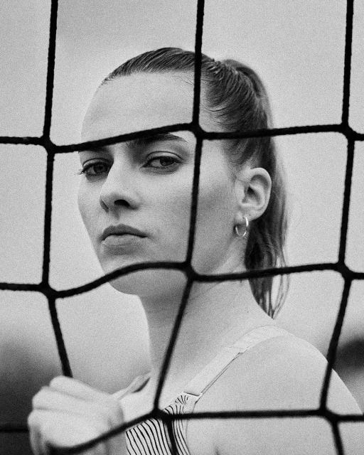 Black and white portrait of a woman with ponytail, seen through a foreground volleyball net, hoop earrings, striped top.