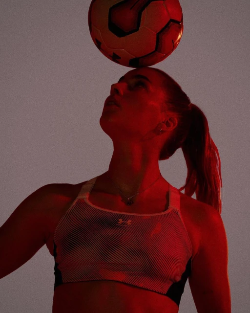 Woman balancing a soccer ball on her head with a red tone, wearing athletic wear against a grey backdrop, showing sport and skill.