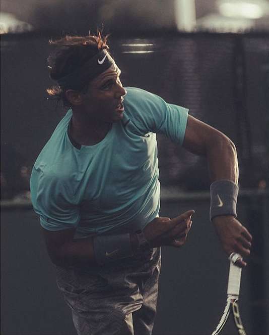 Male athlete in a teal athletic shirt and wristbands, wearing a black Nike headband, holding a tennis racket on an indoor tennis court.