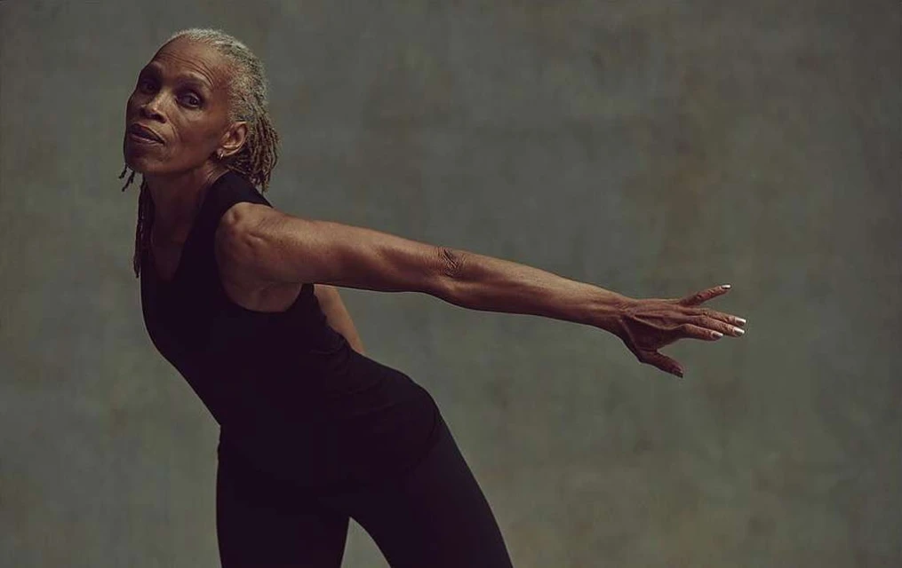 Portrait of an older adult Black woman with grey dreadlocks in a black tank top and black pants performing a modern dance move.
