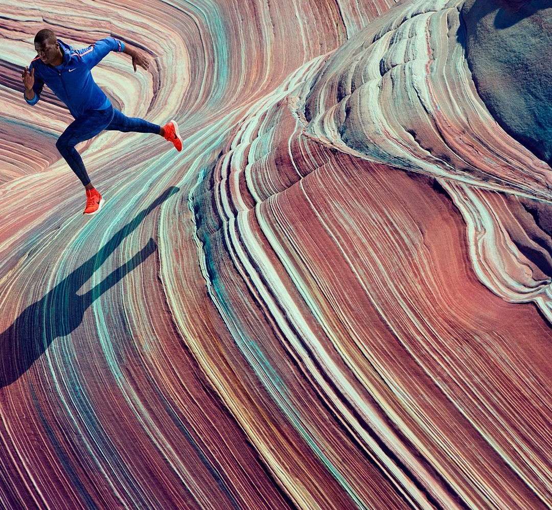 An African American male runner in blue athletic wear sprints across a multicolored, wavy rock formation, casting a long shadow.