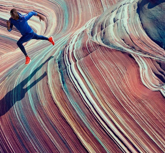 An African American male runner in blue athletic wear sprints across a multicolored, wavy rock formation, casting a long shadow.