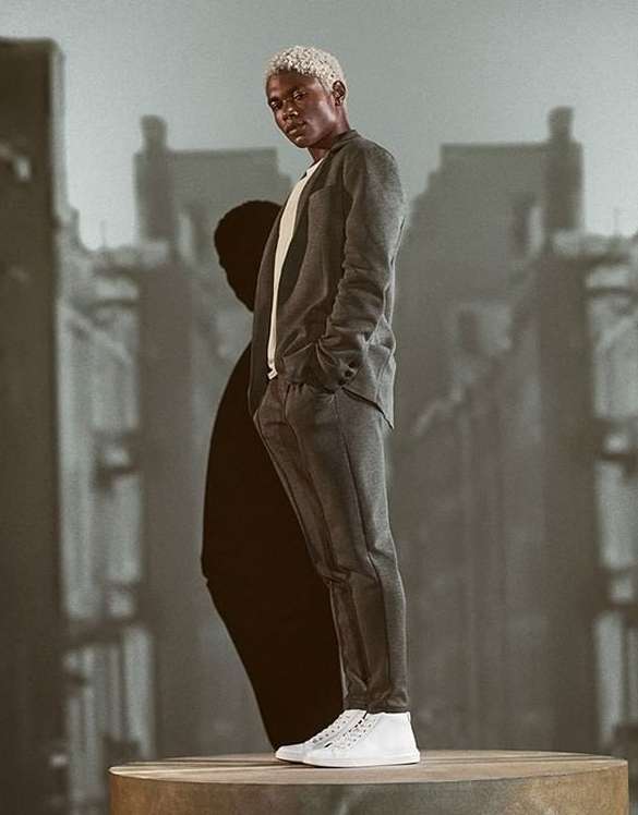 Stylish man with light hair poses in gray suit and white sneakers on a platform against a blurred city backdrop, fashion forward.