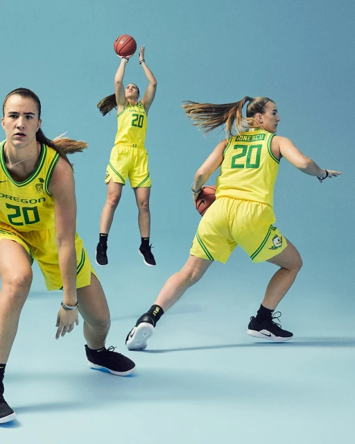 Female basketball player in action, wearing an Oregon Ducks jersey, shooting a ball against a light blue backdrop in a dynamic studio shot.