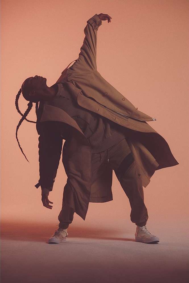 Fashion shot of person in tan coat, pants and shoes posing with arm raised against orange background in studio, full shot.
