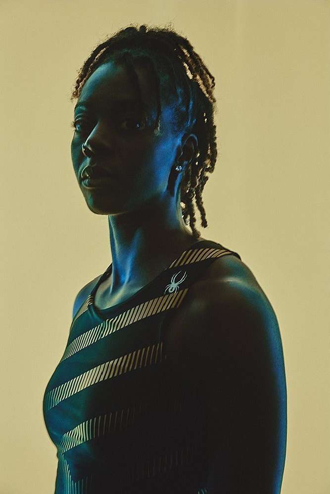 Portrait of athletic, young, Black woman in a striped workout top against a neutral background with strong side lighting.