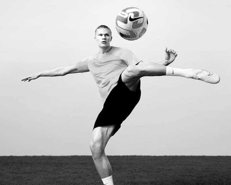 Black and white photo of a man kicking a soccer ball with his right leg, with a grassy field and light grey sky in the background.