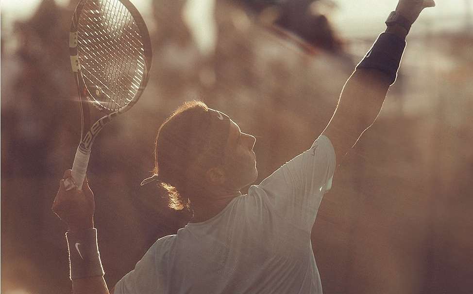 Tennis player serves the ball with tennis racket in hand, wearing wristbands, against a blurred outdoor background in warm lighting.