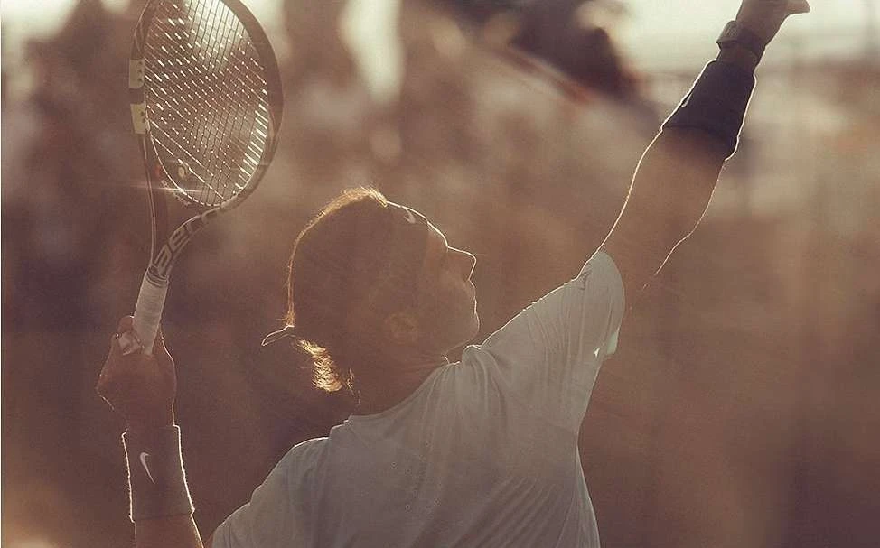 Tennis player serves the ball with tennis racket in hand, wearing wristbands, against a blurred outdoor background in warm lighting.