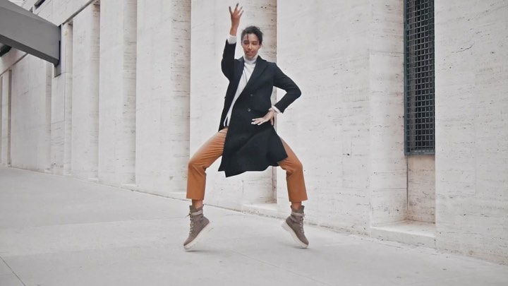 Adult woman in a black coat and tan pants jumps outdoors with her arms raised in front of a wall with architectural features.