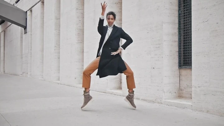 Adult woman in a black coat and tan pants jumps outdoors with her arms raised in front of a wall with architectural features.