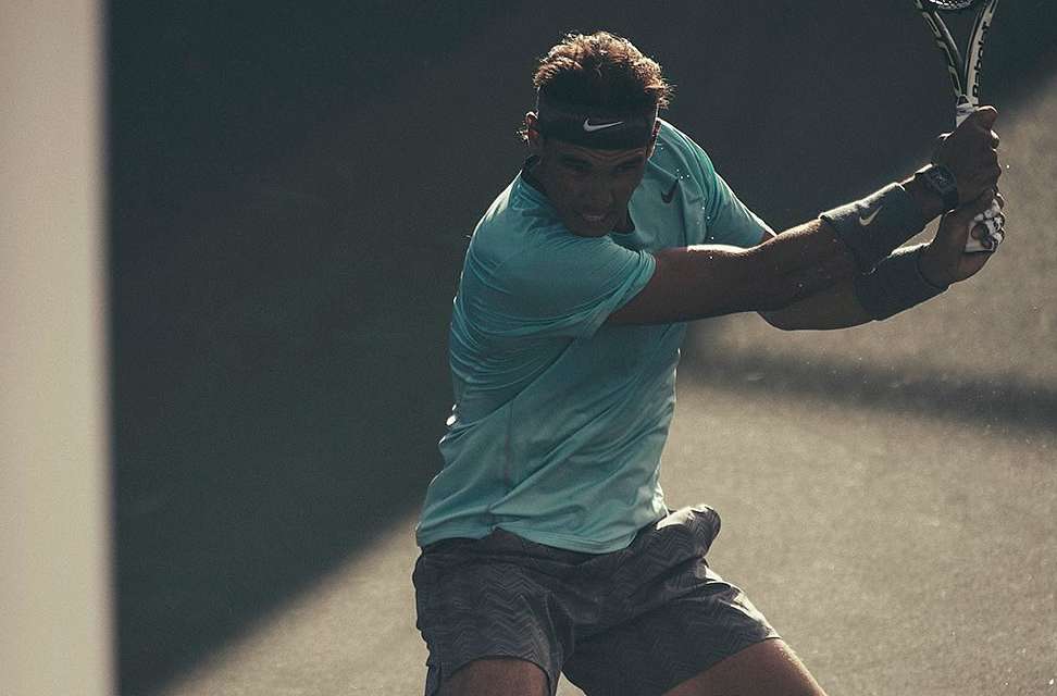 Professional tennis player in action, Rafael Nadal, swinging his racket during a match with a determined facial expression.