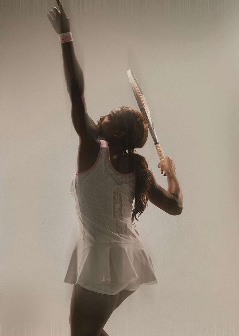An adult female tennis player in a white tennis dress is in mid-swing with a tennis racket held high on a light background in a studio shot.